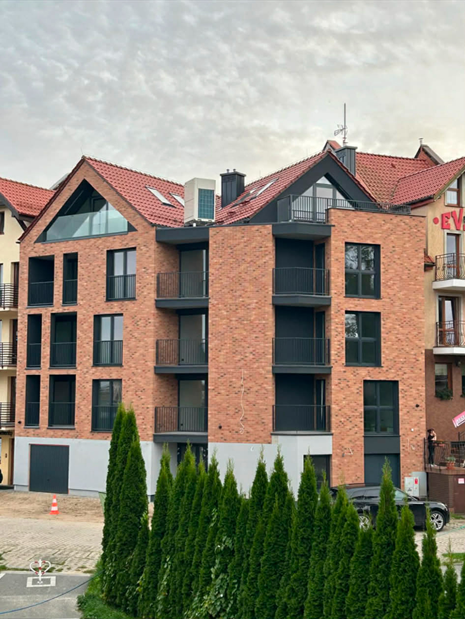 Czteropiętrowy nowy budynek z czerwonej cegły, tarasami i czarnymi balustradami, przed nim szpaler żywotników