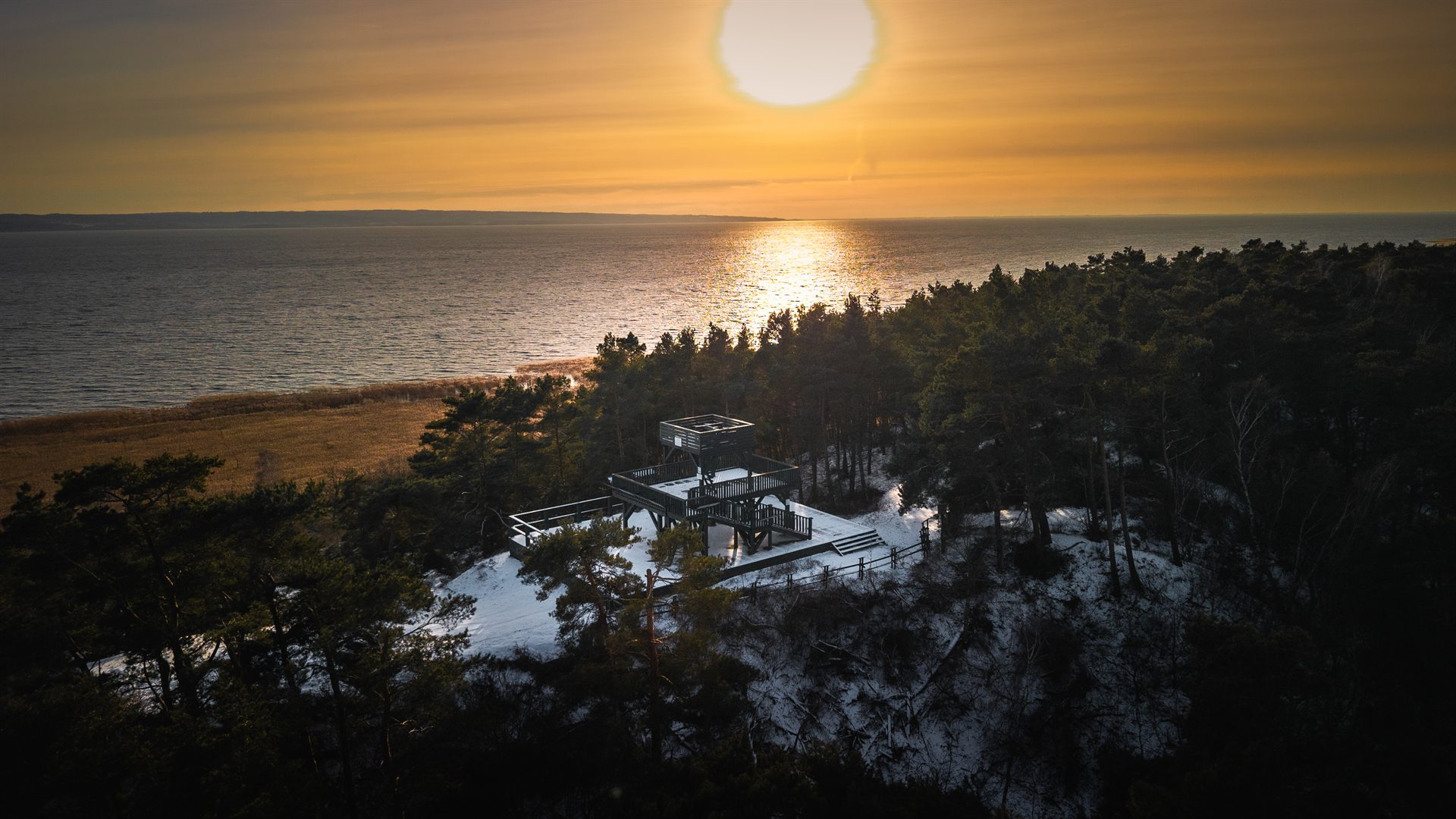 drewniana platforma widokowa na zaśnieżonym klifie otoczona sosnowym lasem z zachodzącym słońcem nad morzem