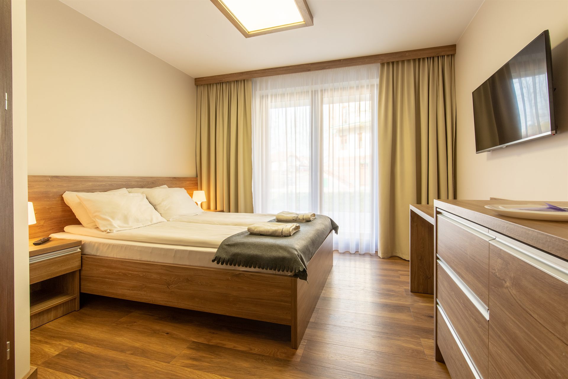 Sypialnia z podwójnym łóżkiem w drewnianej ramie, kremowe pościele, zasłony i telewizor na ścianie