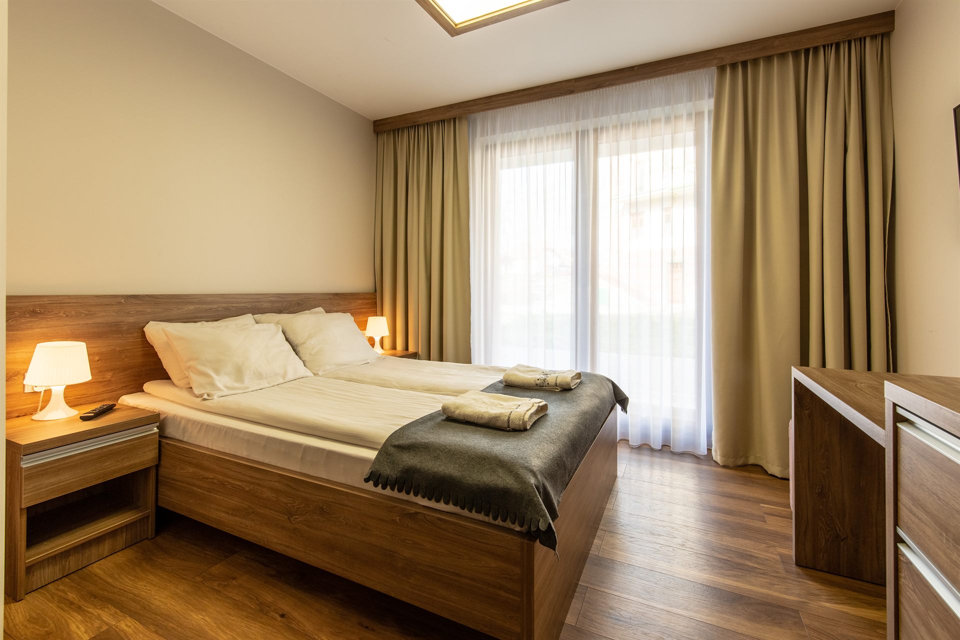 Dwuosobowe łóżko z drewnianą ramą, białą pościelą, ciemnym pledem i złożonymi ręcznikami przy oknie z zasłonami