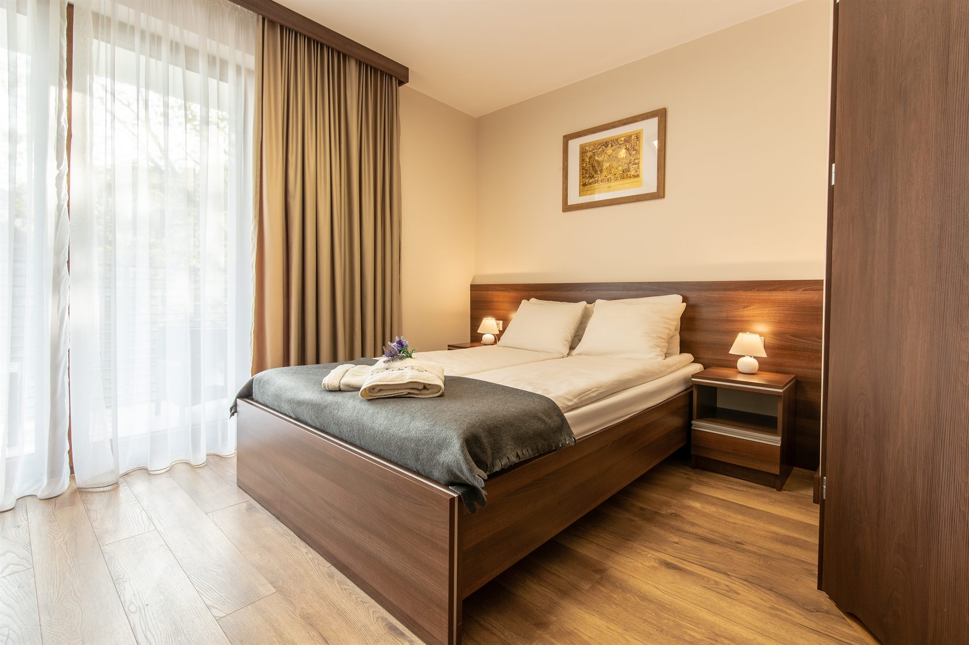 Przytulna sypialnia z drewnianym łóżkiem, jasną pościelą, szarą narzutą i zasłonami oraz stolikiem z lampką