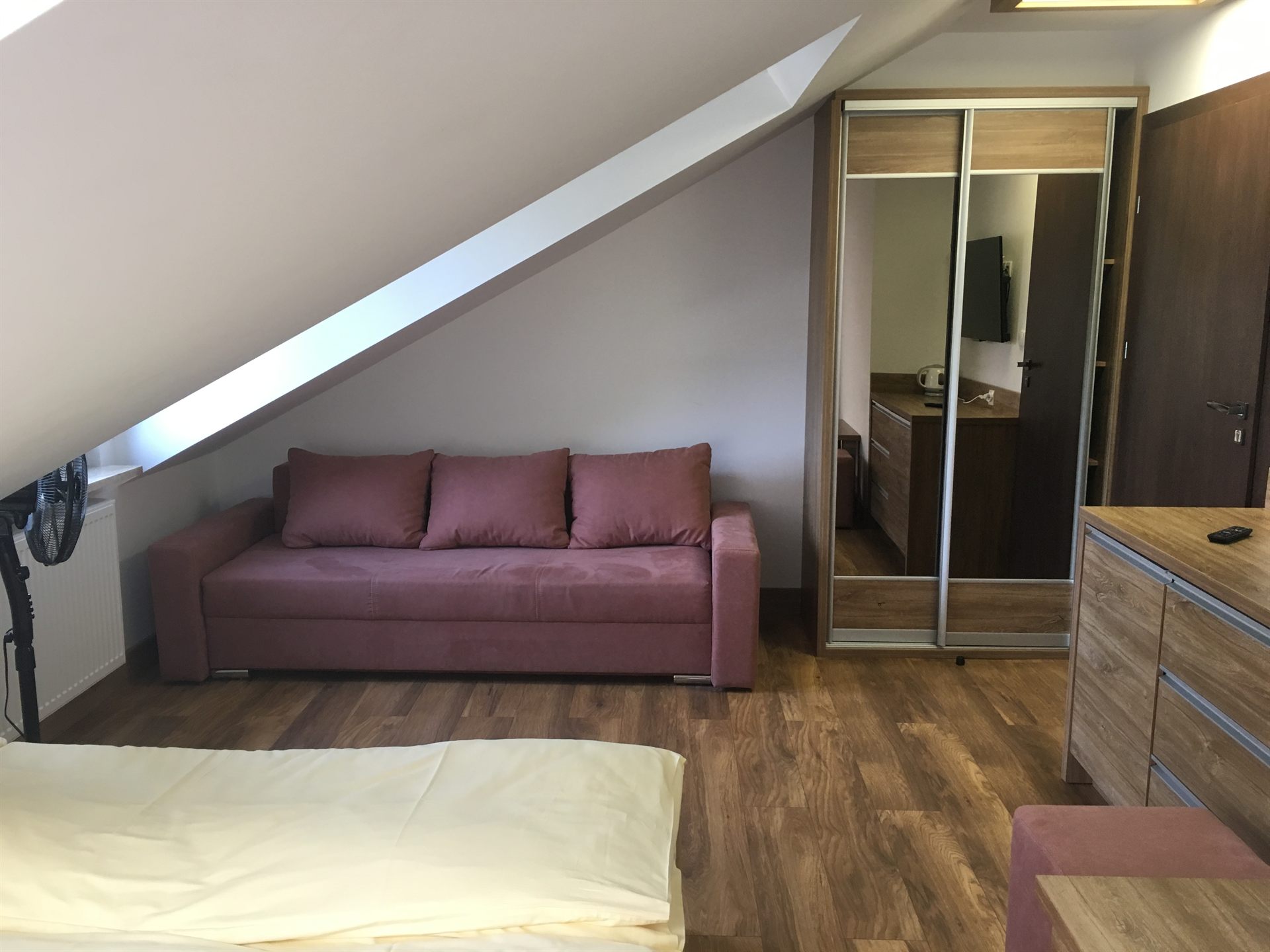 Różowa sofa z trzema poduchami pod skośnym sufitem na poddaszu, po prawej szafa z lustrzanymi drzwiami