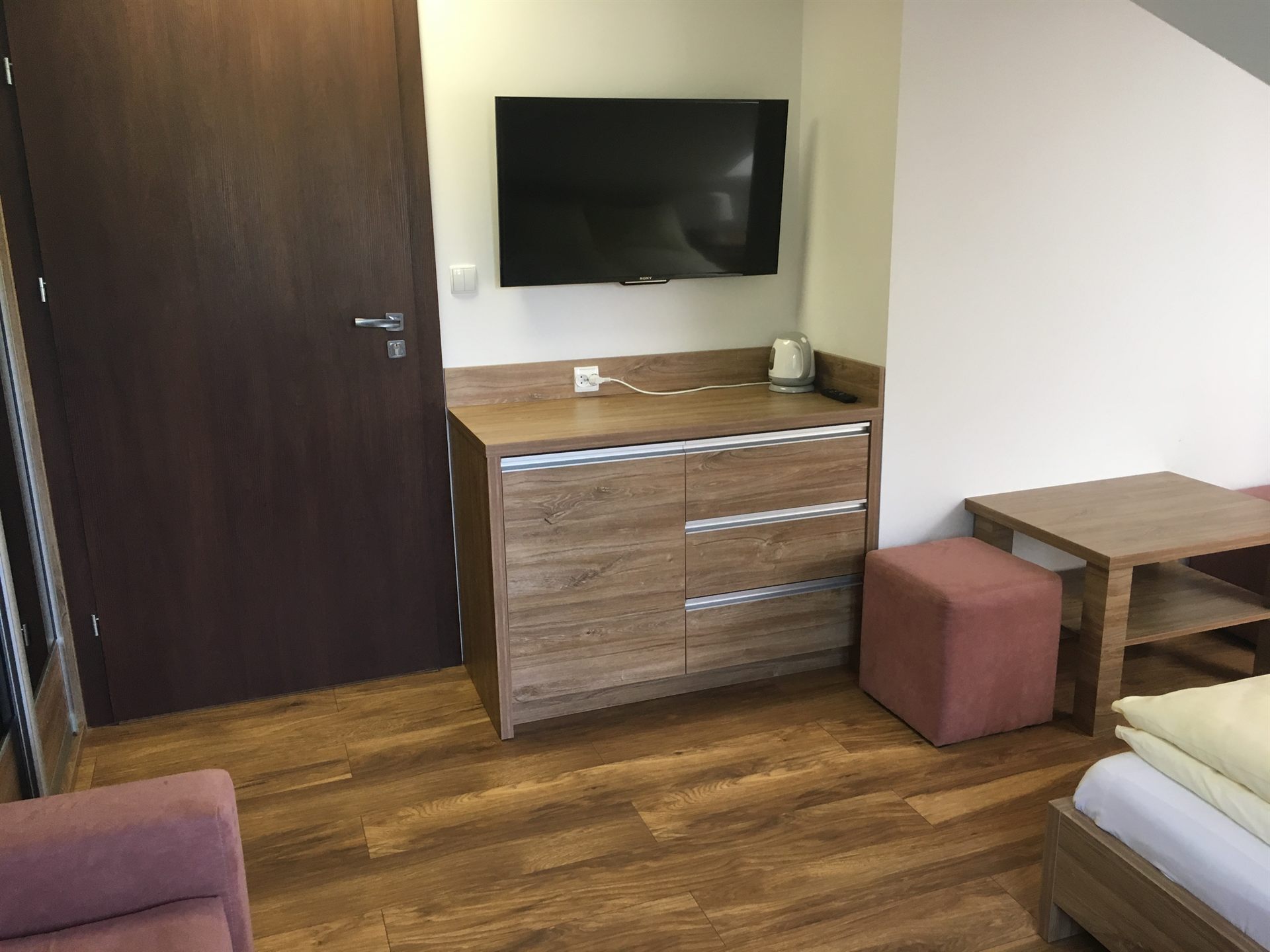 Mały hotelowy kącik z drewnianą komodą, zawieszonym telewizorem, czajnikiem, różowym pufem i stolikiem przy łóżku