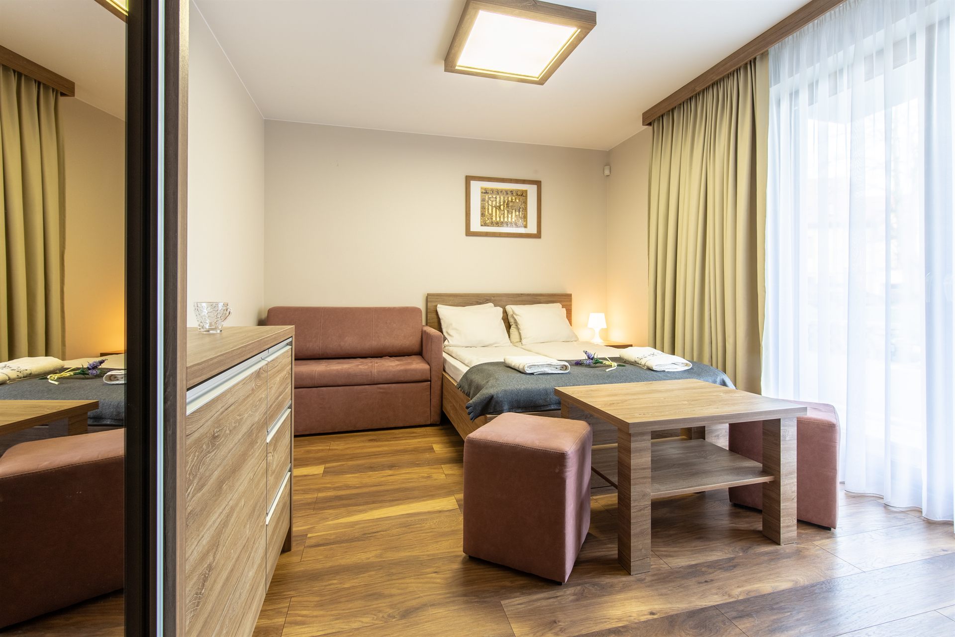 Przytulny pokój z łóżkiem podwójnym, brązową sofą, drewnianym stolikiem i dużym oknem z zasłonami