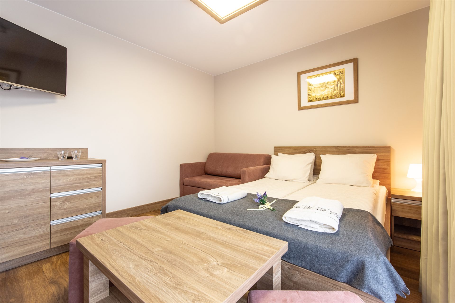 Sypialnia z podwójnym łóżkiem, drewnianymi meblami, szarą narzutą i brązową sofą przy ścianie