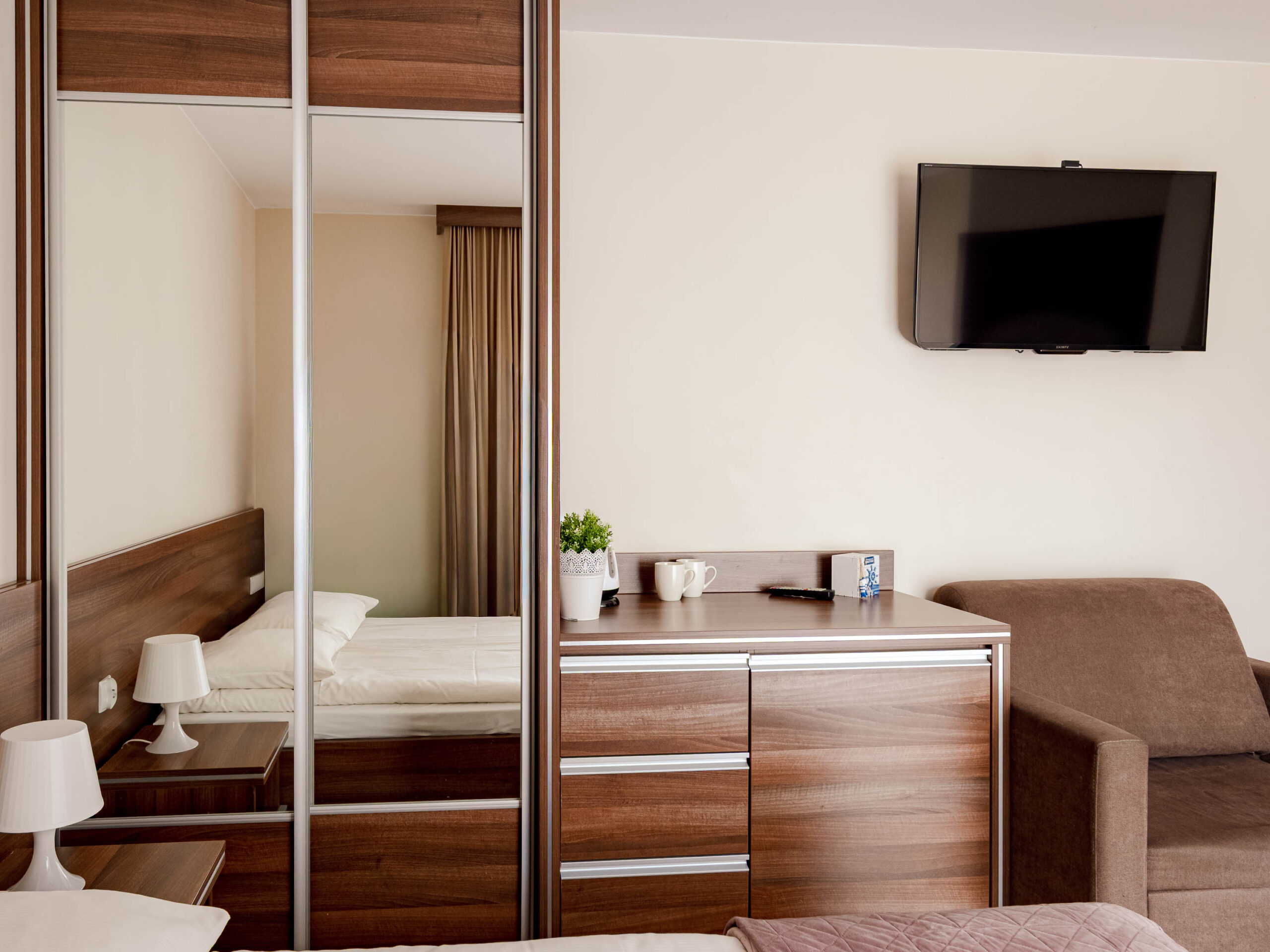 Nowoczesny pokój hotelowy z łóżkiem, szafą z lustrami, komodą, fotelem i telewizorem na ścianie