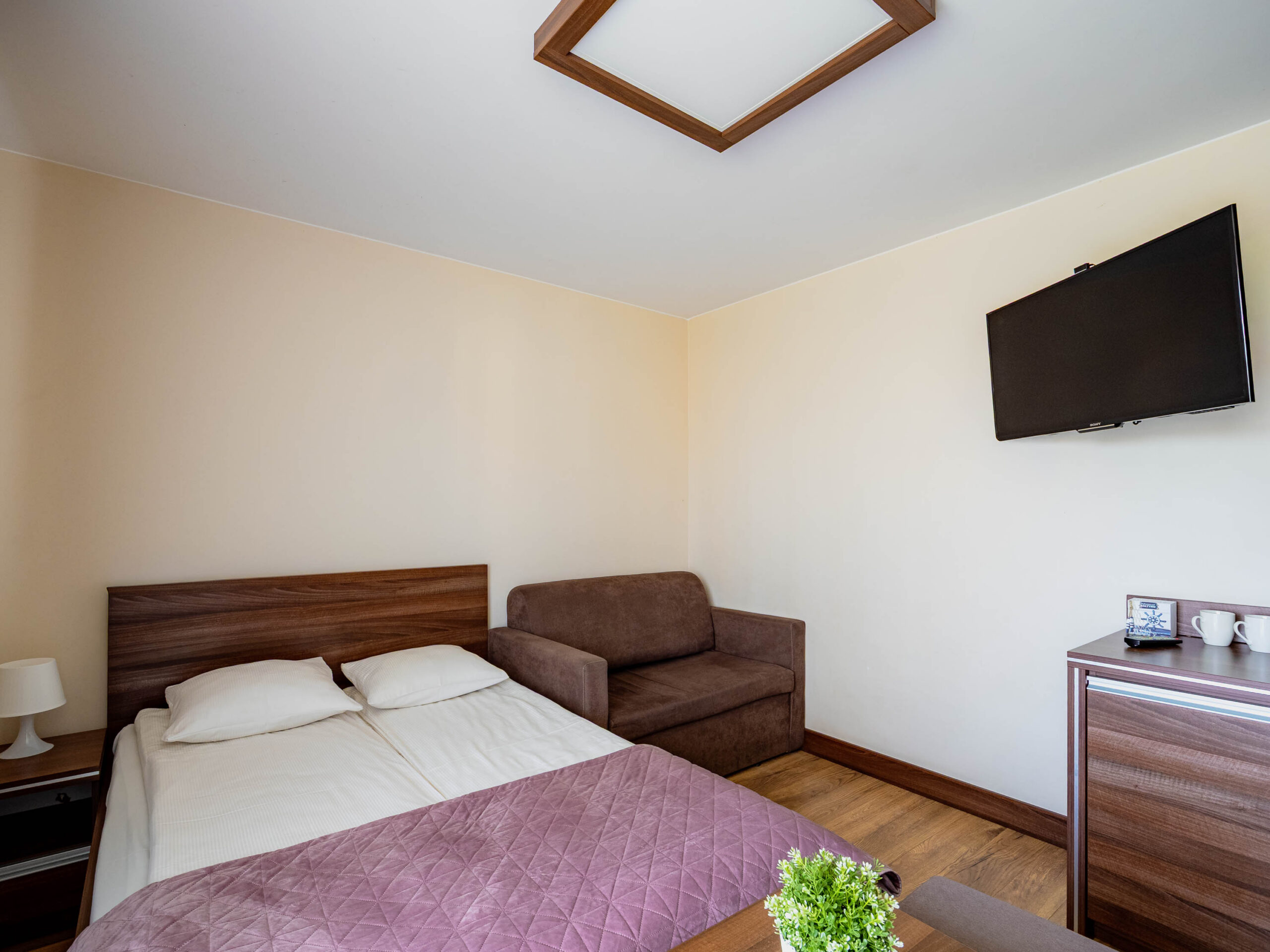 Jasny pokój hotelowy z łóżkiem, fotelem, telewizorem i stolikiem z czajnikiem